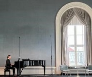 Tanja Huppert plays the folk song in a hall of the Bavarian Academy of Fine Arts. Tanja can be seen far away at a grand piano on the left edge of the picture. The hall looks majestic with high windows in the round arch - like in a castle - and the hall has light blue walls.